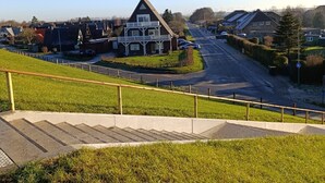 A residential area with a prominent house featuring a balcony, a road with parked cars, and a grassy hill with a staircase. - Holiday Apartment 'Friedrichskoog Spitze' with Private Terrace and Wi-Fi (Friedrichskoog)