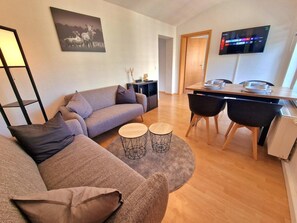 A modern living room with a sofa, coffee table, dining table with chairs, a TV mounted on the wall, and a bookshelf. - Apartment 'Fewo Anne, Großzügige' with Private Terrace, Shared Garden and Wi-Fi (Chemnitz)