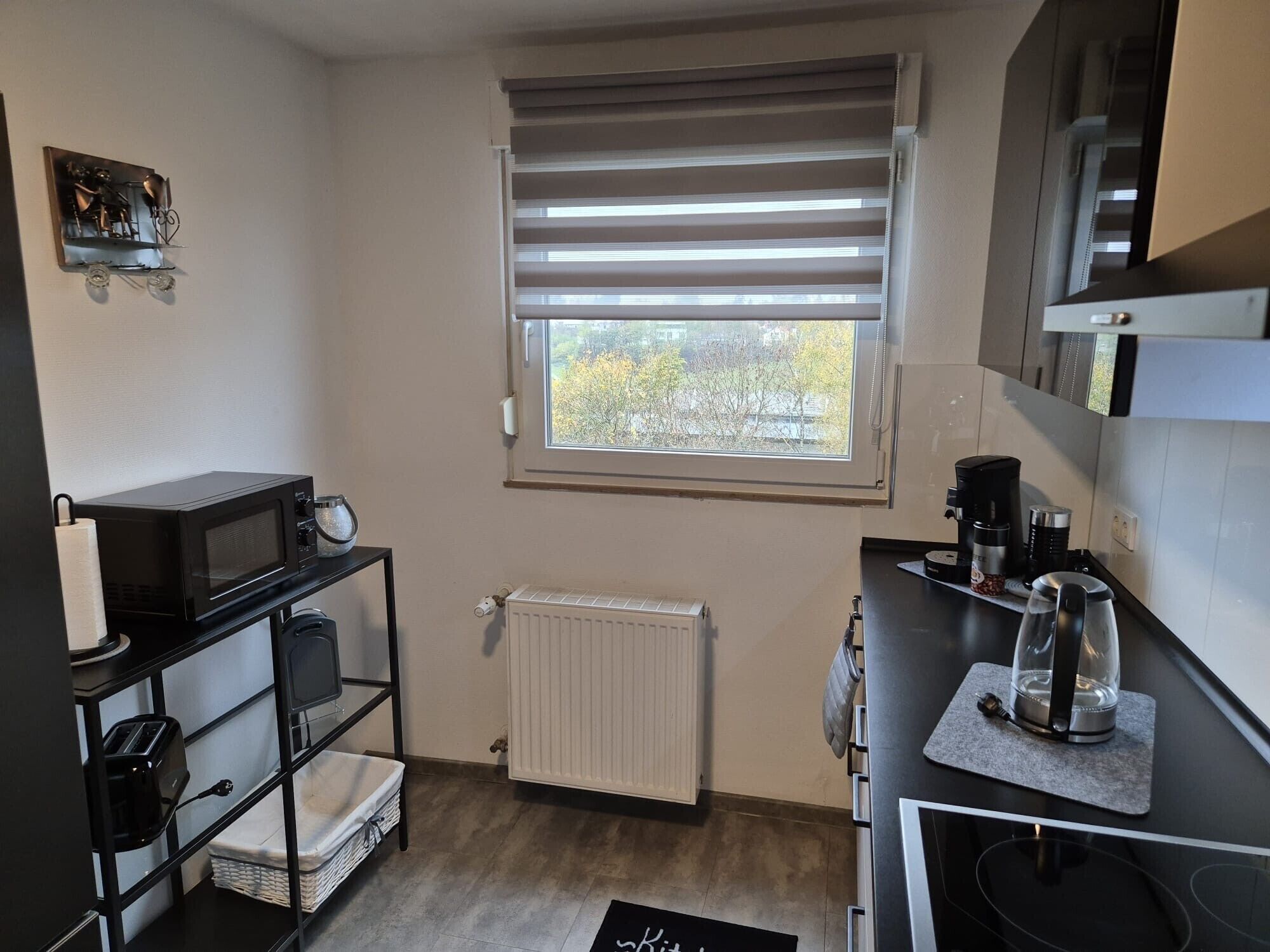 A compact kitchen with a microwave, toaster, and kettle on shelves, a window with blinds, a radiator, and a coffee maker on the counter.