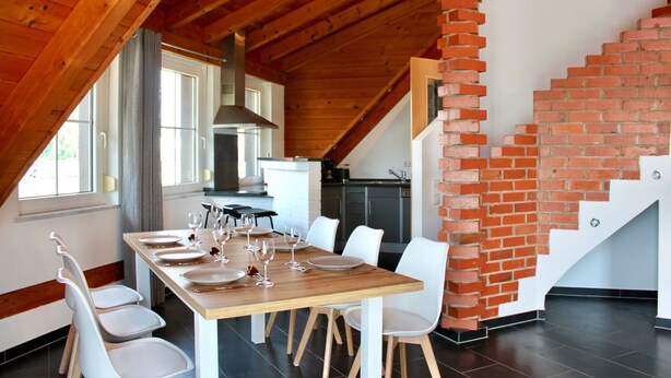 Un comedor con una mesa de madera dispuesta para cuatro personas, sillas blancas y una escalera de ladrillo.