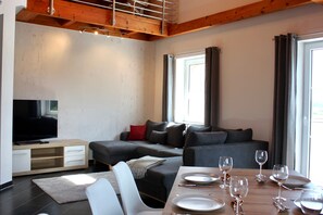 Un salon moderne avec un canapé, un téléviseur, une table à manger pour quatre personnes et une grande fenêtre avec des rideaux.
