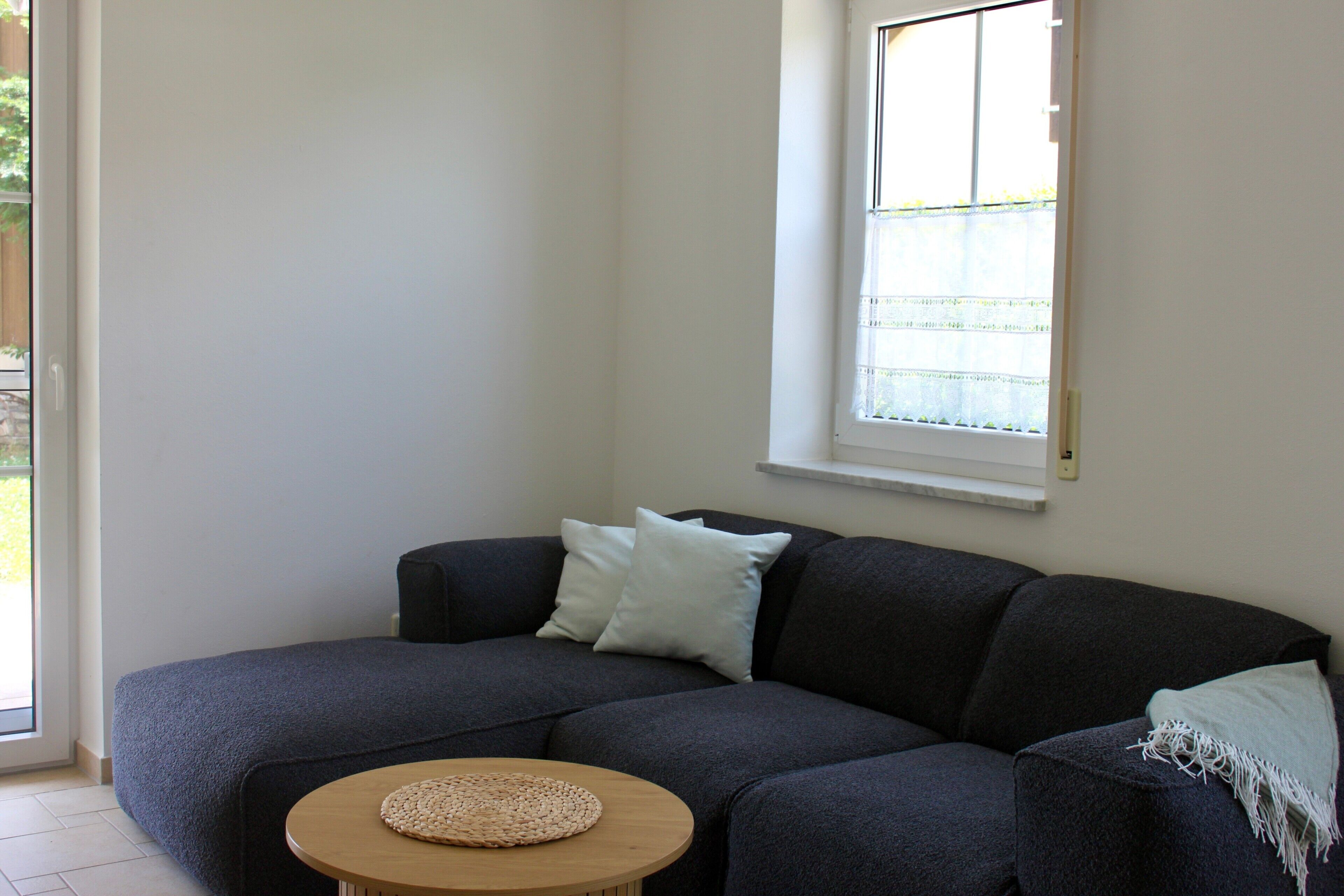 Ein Wohnzimmer mit einem dunkelgrauen Sofa, einem runden Holztisch und einem Fenster mit einem weißen Rollo.