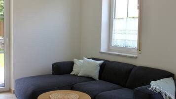Ein Wohnzimmer mit einem dunkelgrauen Sofa, einem runden Holztisch und einem Fenster mit einem weißen Rollo.