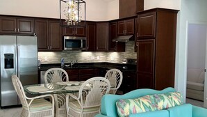 A compact kitchen with dark wooden cabinets, stainless steel appliances, and a glass dining table with wicker chairs.