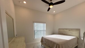 A bedroom with a bed, a ceiling fan, a window with blinds, and a bathroom with a sink and mirror.