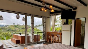 A bedroom with a large bed, a wooden desk with two stools, a TV, and a view of a green landscape. - GLAMPING WITH POOL AND JACUZZI AT GUATAPE  (Antioquia)