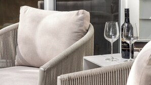 Una silla de mimbre con un cojín, una mesita, dos copas de vino y una botella sobre un balcón.