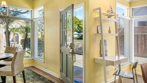 A room with large windows, a door, a small table with chairs, and a shelf with decorative items.