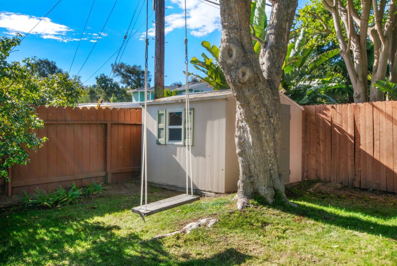 A small, single-story building with a wooden deck, surrounded by a wooden fence and a tree.