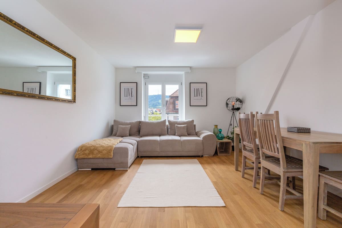 Ein modernes Wohnzimmer mit einer Eckcouch, einem Esstisch mit Stühlen, einem großen Spiegel und gerahmten Bildern an den Wänden.