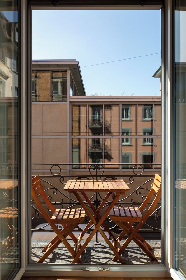 Un balcón con una mesa y sillas de madera, vista a un edificio con múltiples ventanas, y cielo despejado.