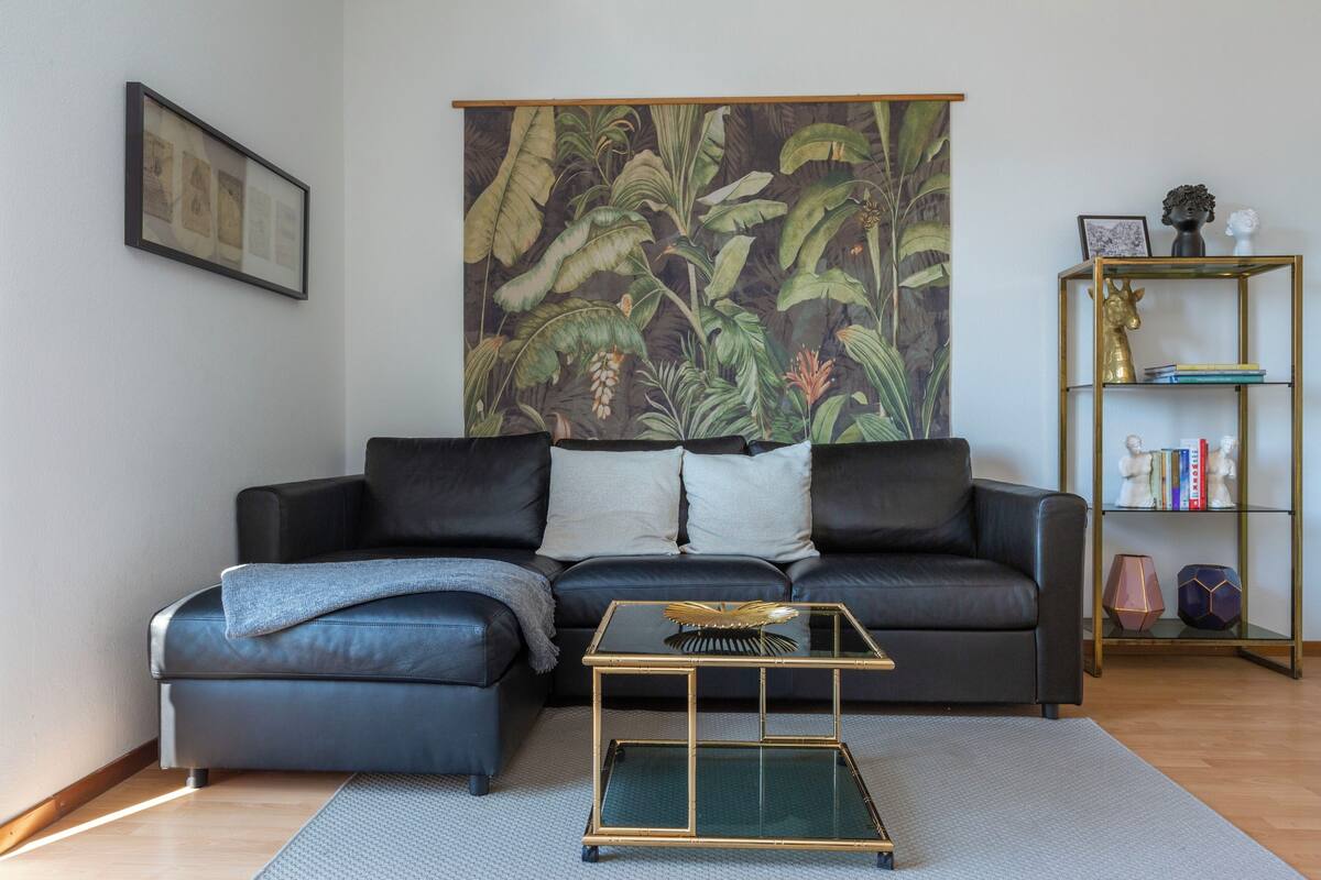 Ein Wohnzimmer mit einem schwarzen Ledersofa, einem Glastisch, einem großen botanischen Wandbild und einem goldgerahmten Bücherregal.