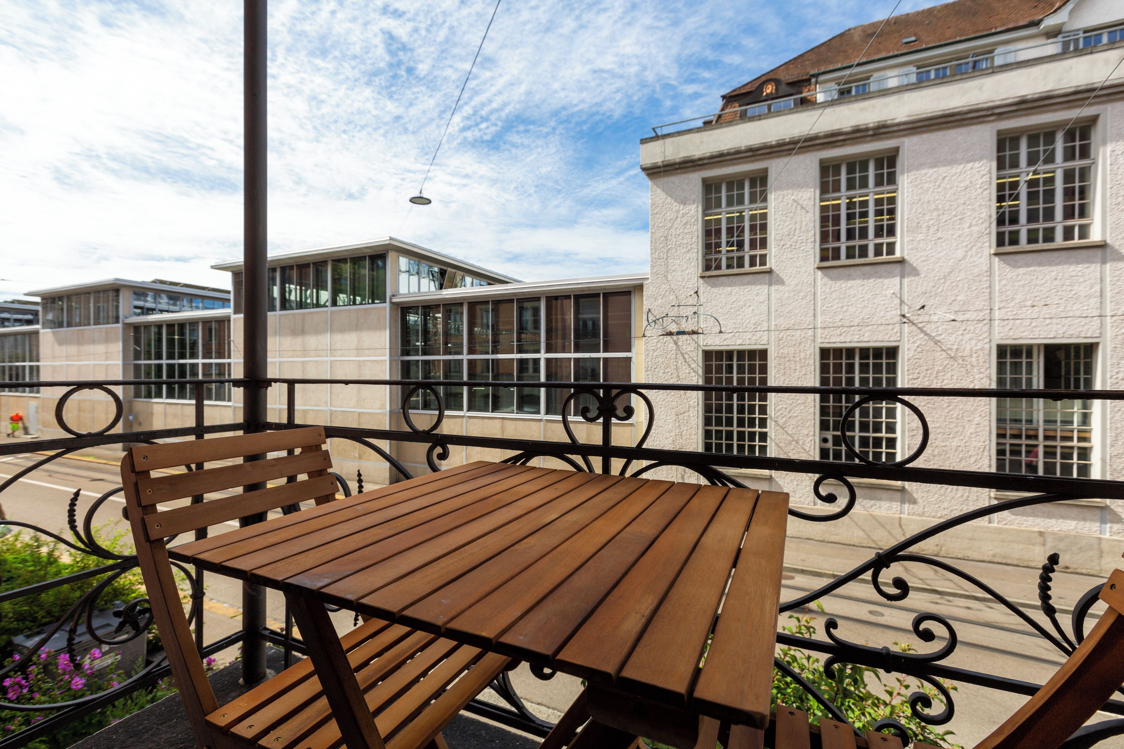 A wooden table and bench on an outdoor balcony with a wrought iron railing, overlooking a street with buildings and a clear sky.