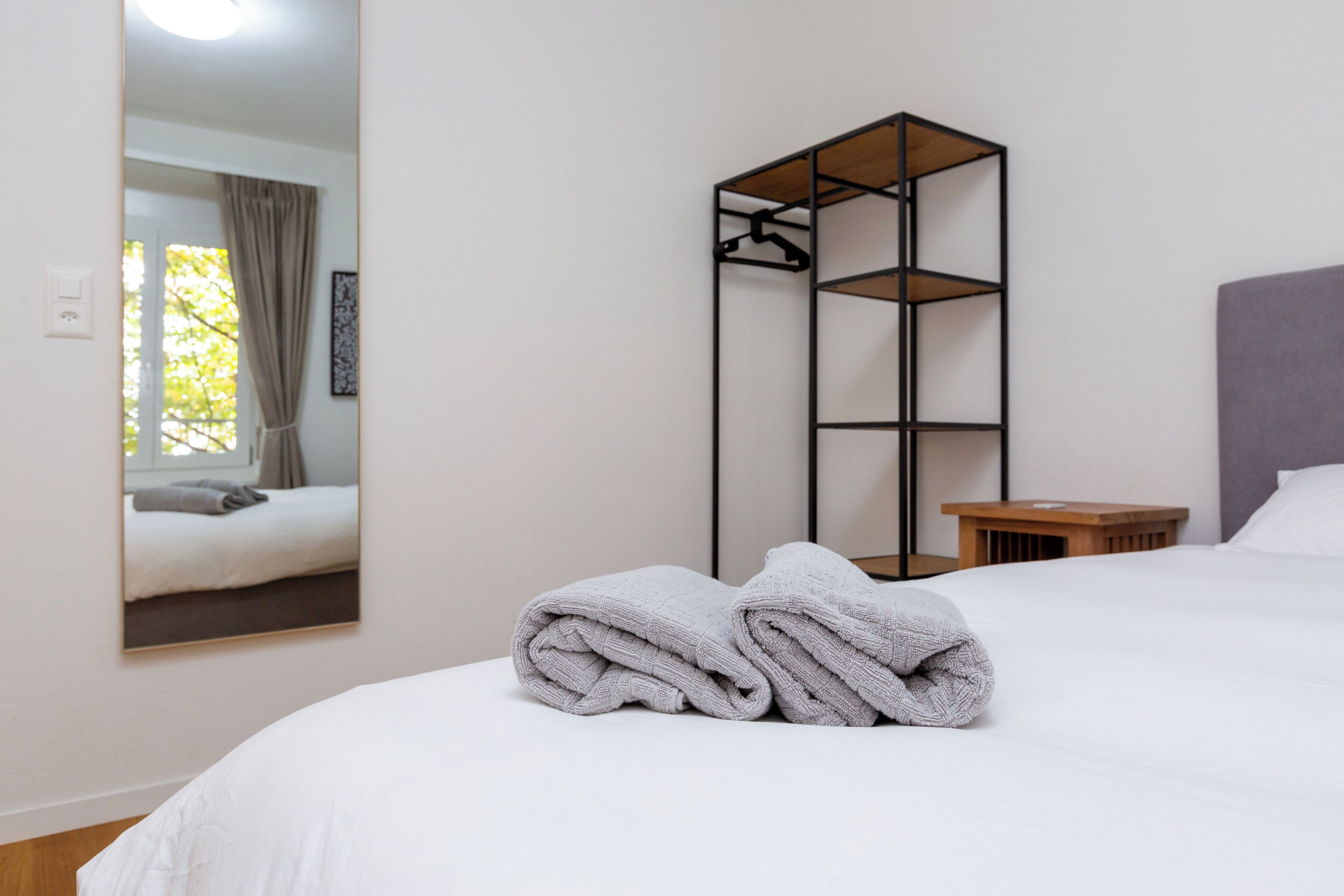 Un lit bien fait, avec une serviette pliée, une table de chevet en bois, une étagère en métal noir et un miroir qui reflète une fenêtre avec des rideaux.
