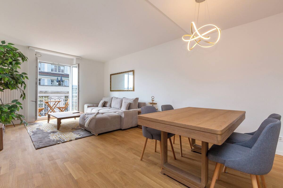 Ein modernes Wohnzimmer mit einem Holzespeisetisch, einem grauen Sofa und einem großen Fenster mit Blick auf Gebäude.