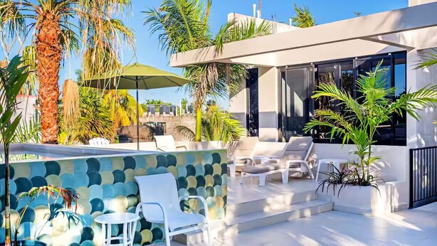 A modern outdoor lounge area with white lounge chairs, a green umbrella, and a decorative wall with a mosaic pattern.