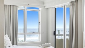 Een slaapkamer met een groot raam met uitzicht op de oceaan, een balkon met een schuifdeur en een net opgemaakt bed met een opgerolde handdoek.