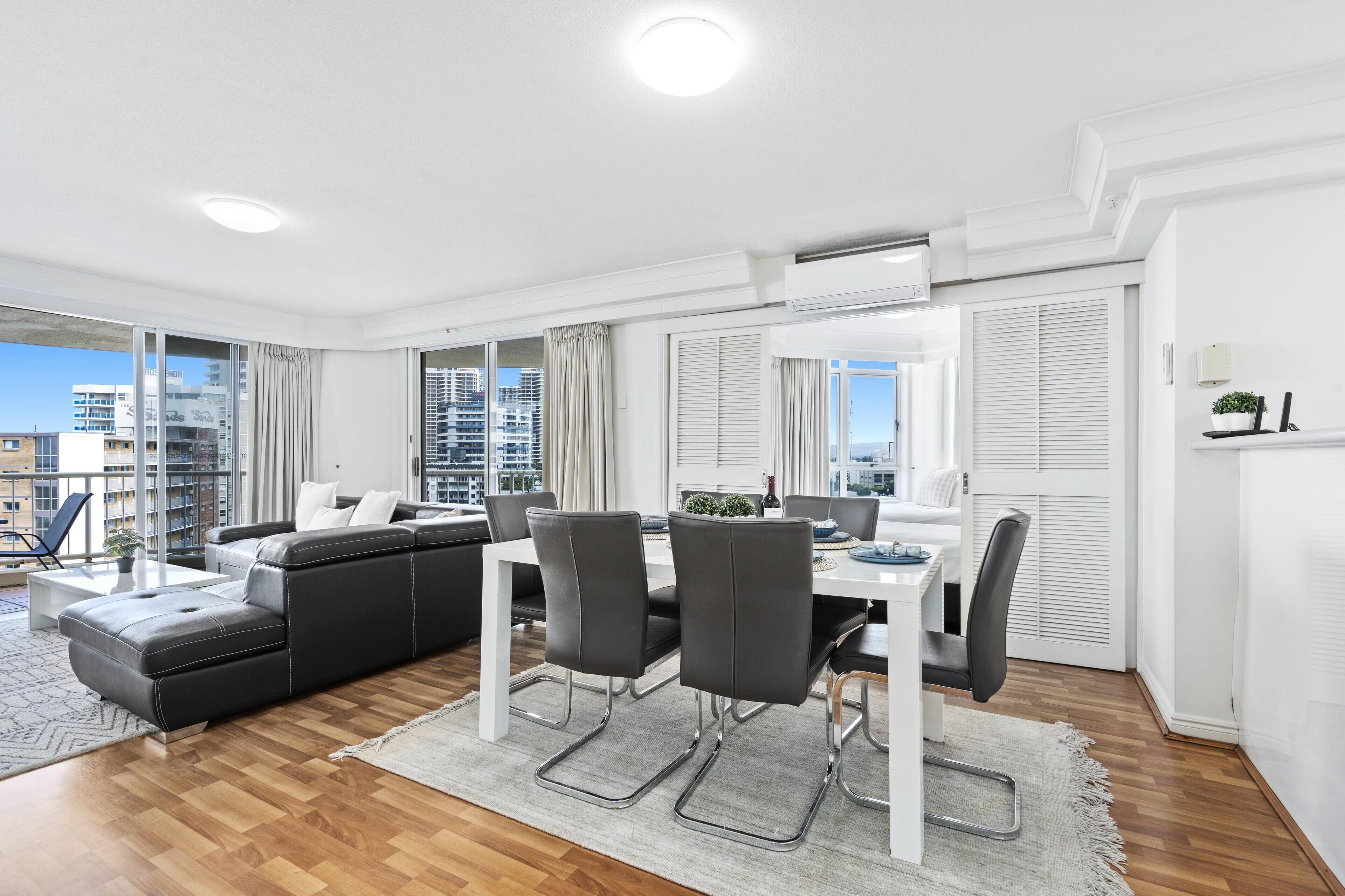 A modern living room with a black leather sofa, dining table, and chairs. Large windows offer a view of the cityscape.