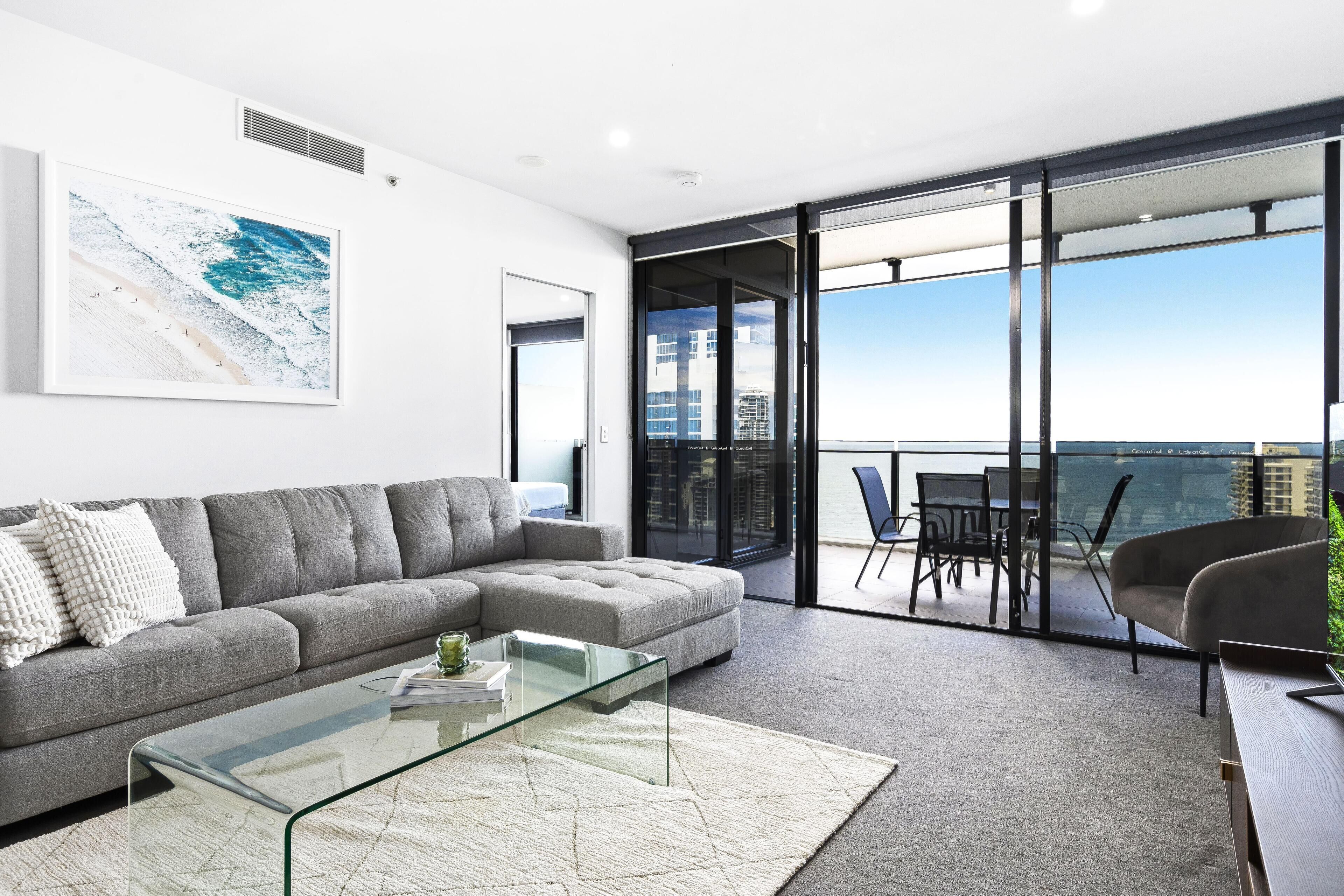 A modern living room with a grey sectional sofa, a glass coffee table, and a large window offering a view of the city.