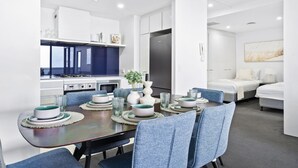 A modern hotel room with a dining area featuring a dark wooden table set with plates, bowls, and glasses. There are blue upholstered chairs, a white sofa, and a bed with white bedding. - 1 Bedroom + Study Ocean View Apartment — Q Stay (COC 1313 - 2N - NB) (Surfers Paradise)
