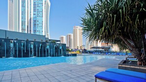 A swimming pool with blue lounge chairs, a waterfall feature, and tall buildings in the background. - 1 Bedroom + Study Ocean View Apartment — Q Stay (COC 1313 - 2N - NB) (Surfers Paradise)