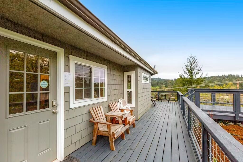 A wooden deck with Adirondack chairs and a table, a door with a window, and a view of trees and hills.