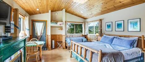 Um quarto aconchegante com teto de madeira, cama com roupa de cama azul, uma mesinha com cadeiras e uma TV em um armário de madeira.