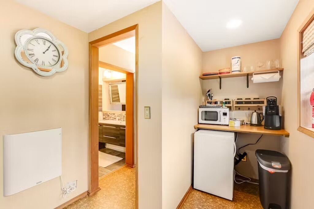 A compact kitchen with a microwave, coffee maker, and a small refrigerator. A clock is mounted on the wall, and there is a trash bin to the right.