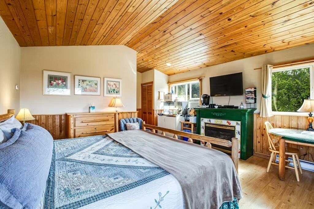 A cozy bedroom with a large bed, a wooden ceiling, a kitchen area with a fireplace, and a dining table.