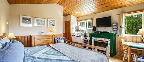 A cozy bedroom with a large bed, a wooden ceiling, a kitchen area with a fireplace, and a dining table.