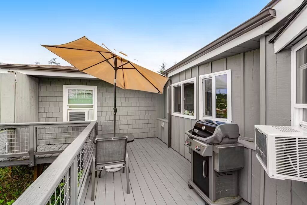 En terrasse med grill og parasol, omgivet af træterrasse og en grå bygning.