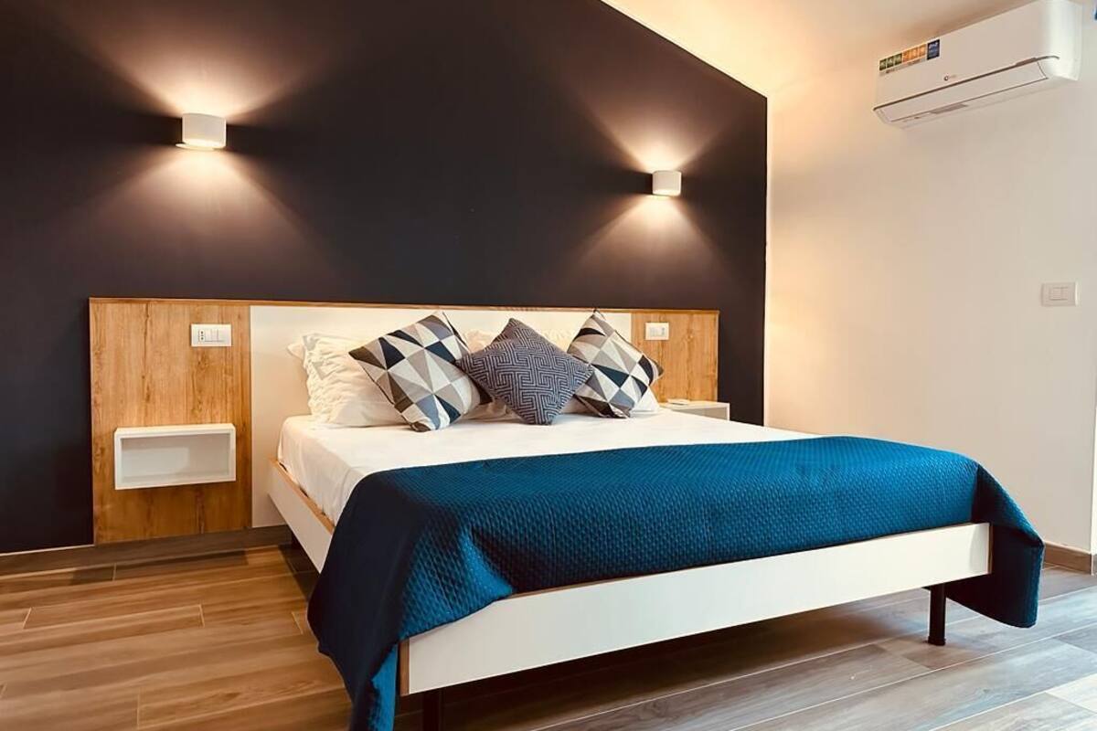 Un dormitorio moderno con cabecera de madera, cama blanca y manta azul. Hay dos mesitas de noche y un aire acondicionado montado en la pared.