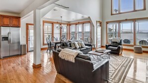 A spacious living area with a large dark sofa, a coffee table, and a dining table with chairs. There is a view of the ocean through large windows.