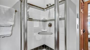 A shower with a glass door, a white tiled wall with a black and green border, and a wooden door to the right.