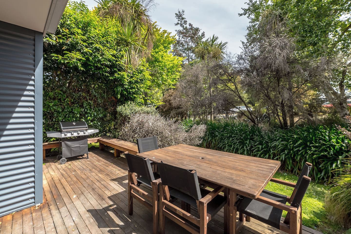 An outdoor patio with a wooden table and chairs, a barbecue grill, and a variety of greenery.