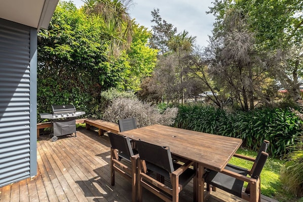 An outdoor patio with a wooden table and chairs, a barbecue grill, and a variety of greenery.