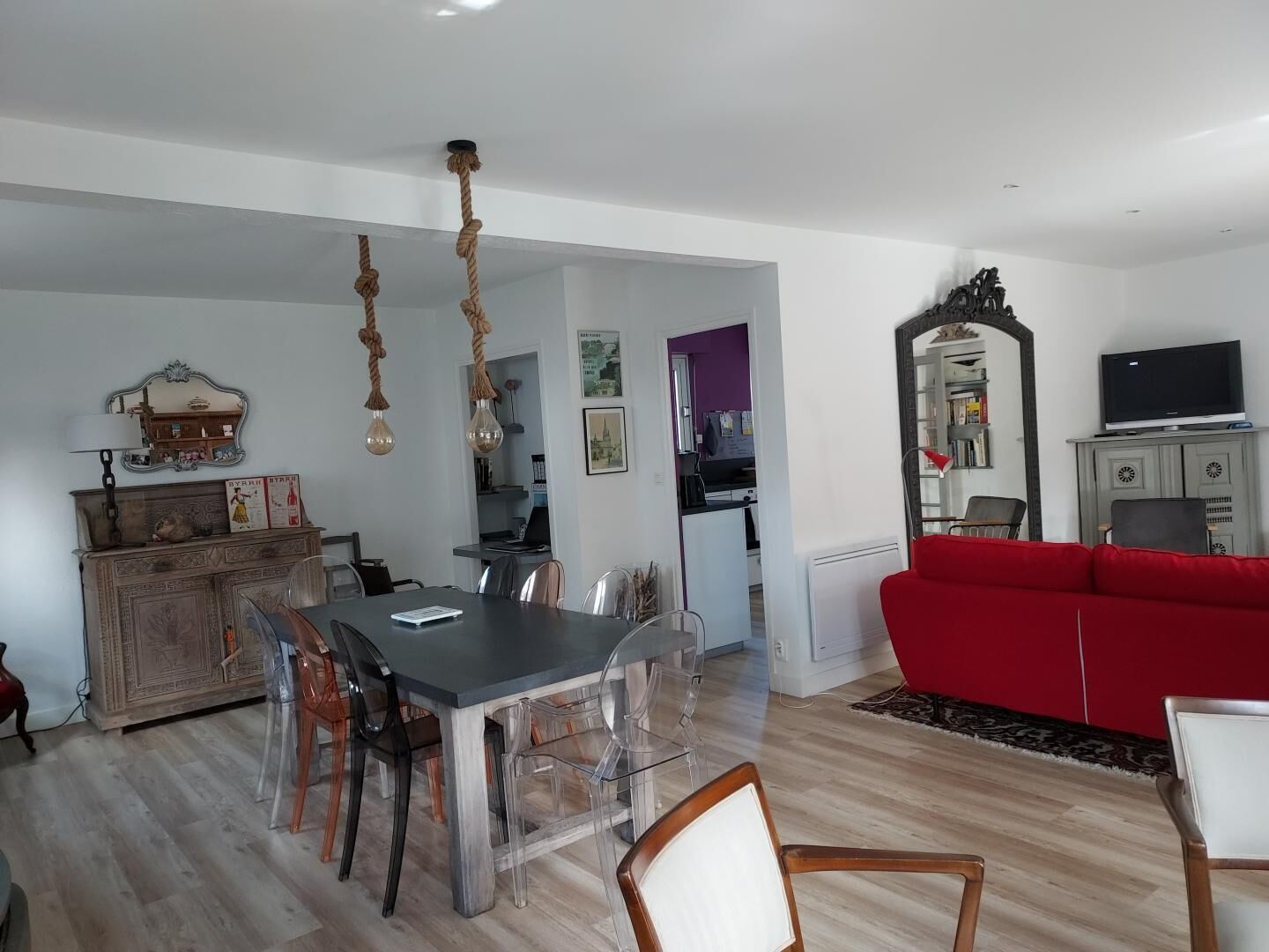 A dining area with a table and chairs, a red sofa, a TV, and a kitchen in the background.