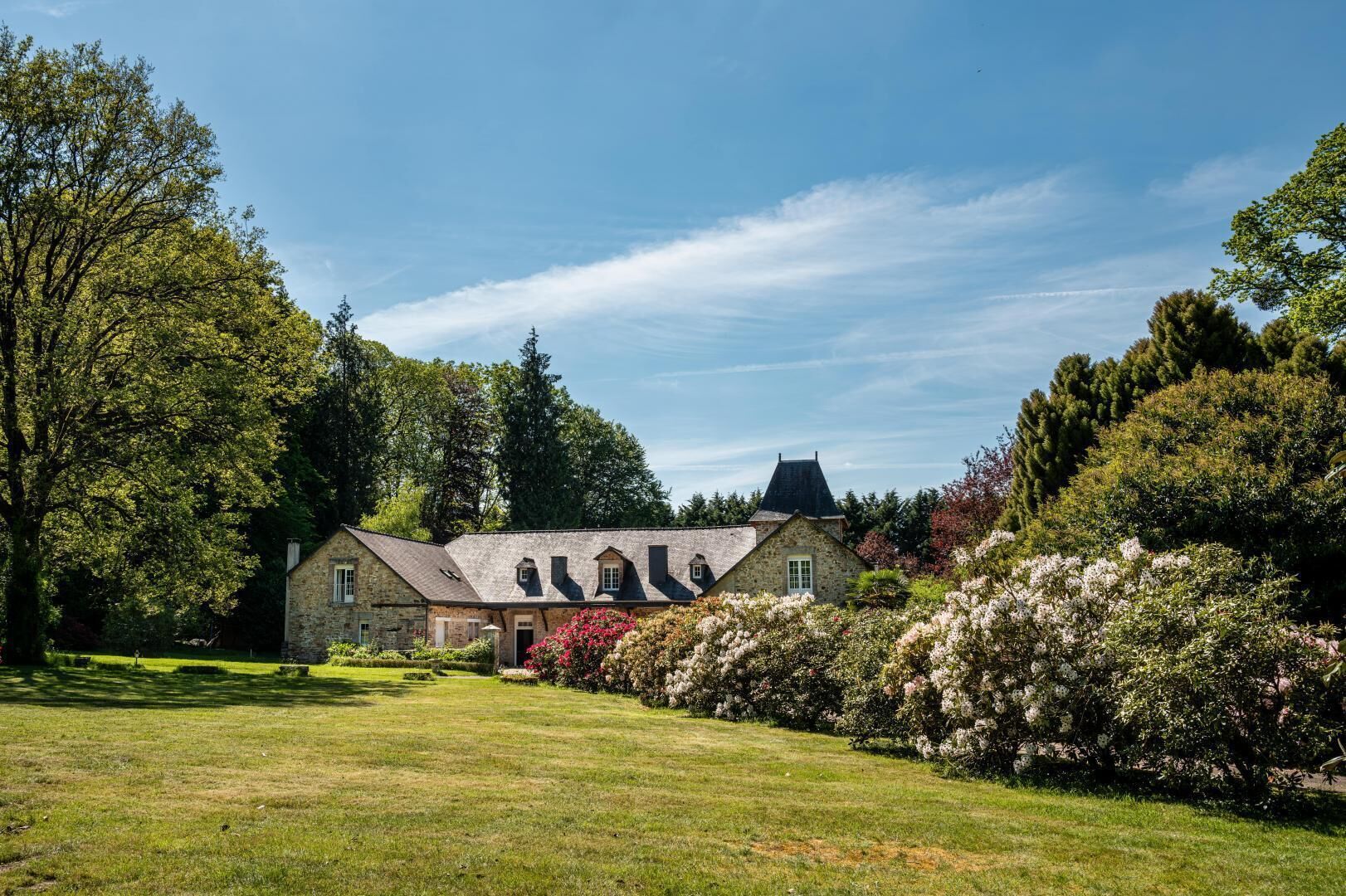 Un bâtiment en pierre doté d’un toit à pignon, entouré d’une végétation luxuriante et de buissons en fleurs.