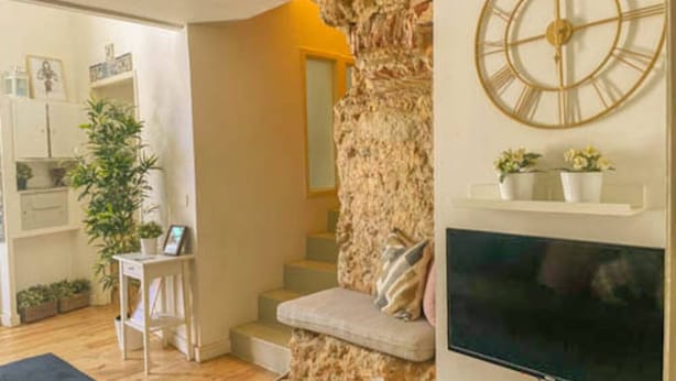 Una sala de estar con una pared de acento de piedra, un reloj de pared, un televisor y una escalera que lleva a otra habitación.