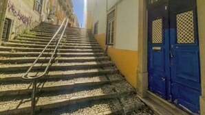 Unos escalones de piedra conducen a un edificio con una puerta azul, barandilla de metal y cielo despejado.
