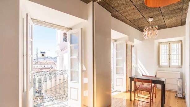 A bright room with wooden floors, a dining table with chairs, a balcony with a view of buildings, and a ceiling with exposed beams.