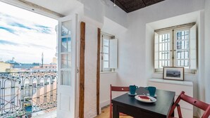 A room with a balcony, a table with chairs, and a window with a picture frame.