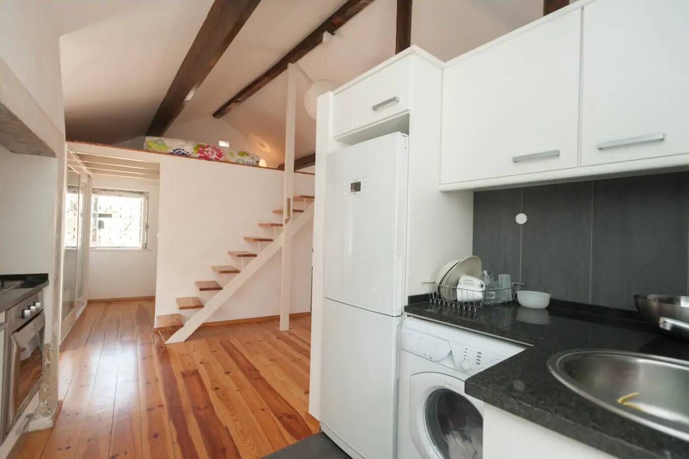 Une cuisine avec un plancher en bois, des armoires blanches, un lave-linge encastré et un comptoir noir.