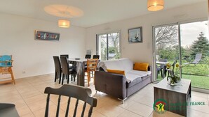 A modern living room with a sofa, dining table, chairs, and large windows offering a view of the outdoors.