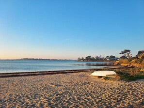 Una playa con un banco blanco, cielo despejado y una costa distante con edificios y árboles.