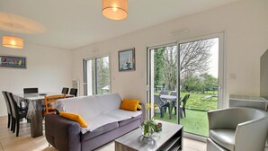 A modern living room with a sofa, coffee table, and dining area. There is a large sliding glass door leading to a garden.