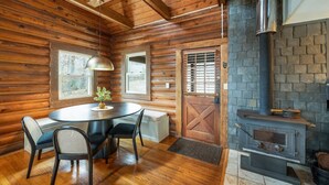 Interior de una cabaña de troncos con una mesa redonda de comedor, sillas, una chimenea y un ventanal grande.