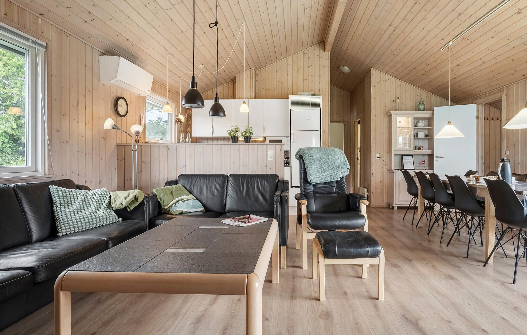 A modern interior with a wooden ceiling, a black leather sofa, a dining area with black chairs, and a kitchen with white cabinets.