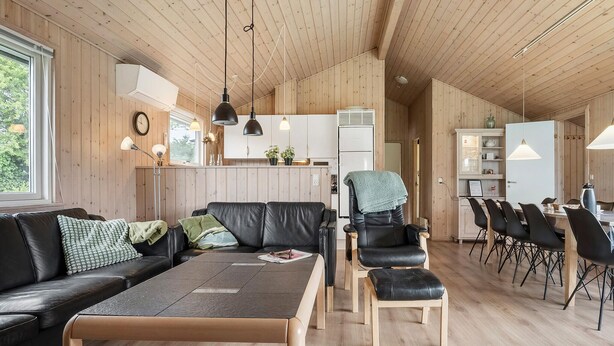 Un intérieur moderne avec un plafond en bois, un canapé en cuir noir, un coin repas avec des chaises noires et une cuisine avec des meubles blancs.