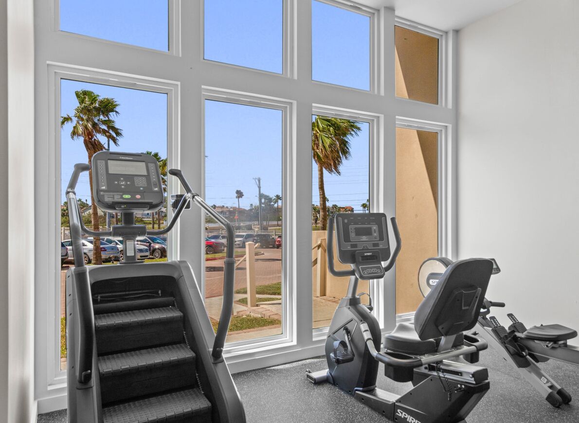 Ein Fitnessstudio mit Laufbändern und einem Crosstrainer, große Fenster mit Blick auf Palmen und einen Parkplatz.